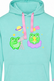 A mint green pullover hoodie featuring a cute cartoon graphic of two green frogs with pink and yellow space-themed accessories, with matching graphics printed on both sleeves.