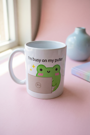 A white ceramic coffee mug with a cute green cartoon frog holding a laptop and the text 'I'm busy on my puter', placed on a pink surface near a window with a small vase in the background.