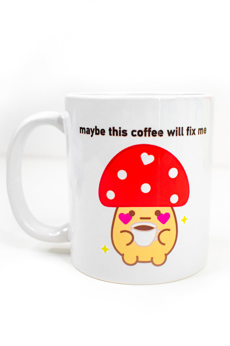A white ceramic mug with a mushroom friend design and the text 'maybe this coffee will fix me' on it, displayed on a brown surface with a floral arrangement in the background.