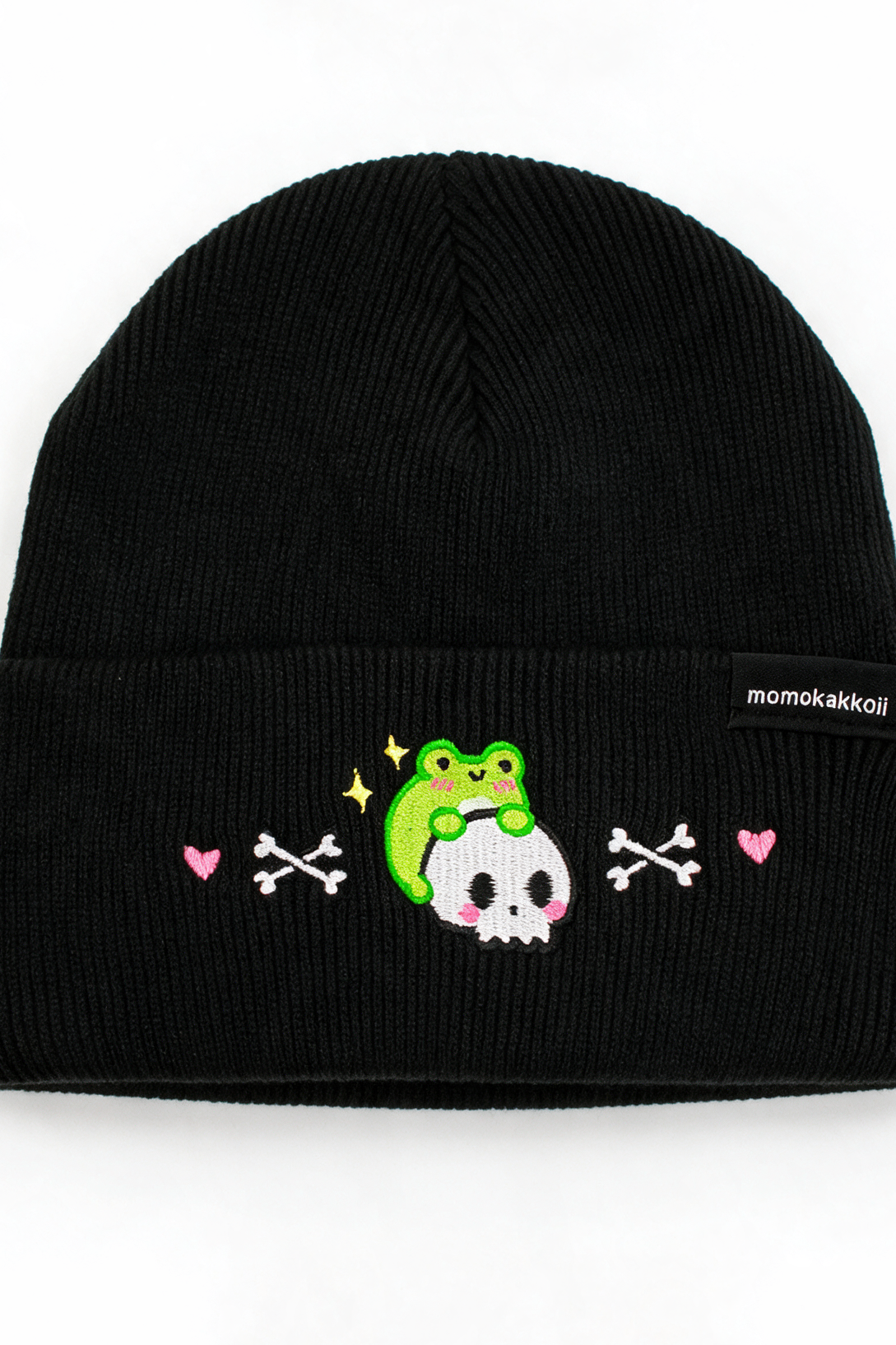 A black ribbed knit beanie hat featuring a cute embroidered design on the front cuff. The design shows a green frog sitting on a white skull, flanked by pink hearts and white crossed bones.