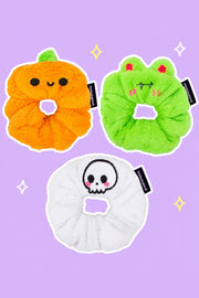 Spooky Scrunchie Trio – Halloween Bundle: one orange Patch the Pumpkin scrunchie with cute round black eyes and curved smile (NOT jack-o-lantern), one lime green Vampire Albert scrunchie with red eyes and fangs, one white Cute Skull scrunchie, on a simple pastel background with white outlines and small sparkles.