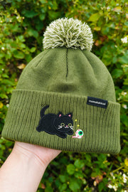 An olive green ribbed knit beanie with a fluffy pom-pom, held up by a hand against a background of green foliage. The beanie features an embroidered black cat wearing round glasses and a small green and white mushroom, with a black brand tag reading 'momakakeo' on the side.