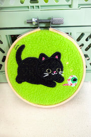A circular embroidery hoop containing bright green felt with an embroidered black cat and a small colorful fish, photographed against a pale green crate background.
