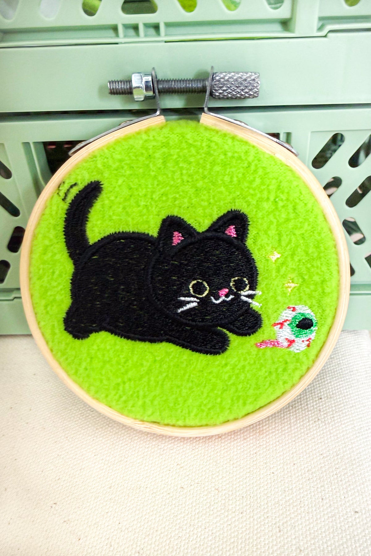 A circular embroidery hoop containing bright green felt with an embroidered black cat and a small colorful fish, photographed against a pale green crate background.