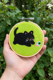 A hand holds a circular embroidery hoop featuring a black cat and a small green-and-white fish on a bright green felt background, with a blurred green garden behind it.