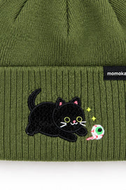 Close-up of an olive green ribbed knit beanie with a black embroidered cat patch and a small black brand tag reading 'momoka' on the cuff.