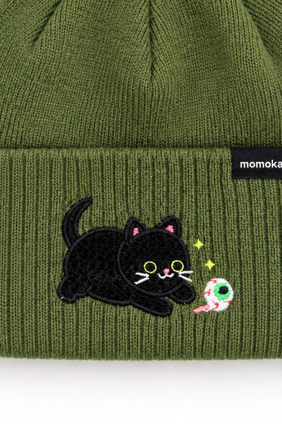 Close-up of an olive green ribbed knit beanie with a black embroidered cat patch and a small black brand tag reading 'momoka' on the cuff.