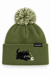 An olive green knit beanie with a pom-pom on top and a folded cuff featuring an embroidered black cat and a small colorful moon, with a small black brand tag reading 'momakakoi' on the side.