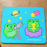 A square mouse pad with rounded corners featuring a cute cartoon design of two green frogs in a space theme, placed on a wooden table. The left frog holds a silver telescope, while the right frog sits on a purple cloud with a yellow star.