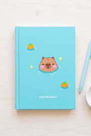 A light blue hardcover notebook with a cute cartoon capybara in a pool and small orange illustrations, styled on a light wood surface next to a matching light blue pen and a small white dish. The brand name 'momokakkoii' is printed at the bottom.