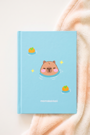 A light blue hardcover notebook with a cute cartoon capybara swimming in water, surrounded by small oranges and stars. The notebook is placed on a soft peach-colored blanket on a light beige surface.
